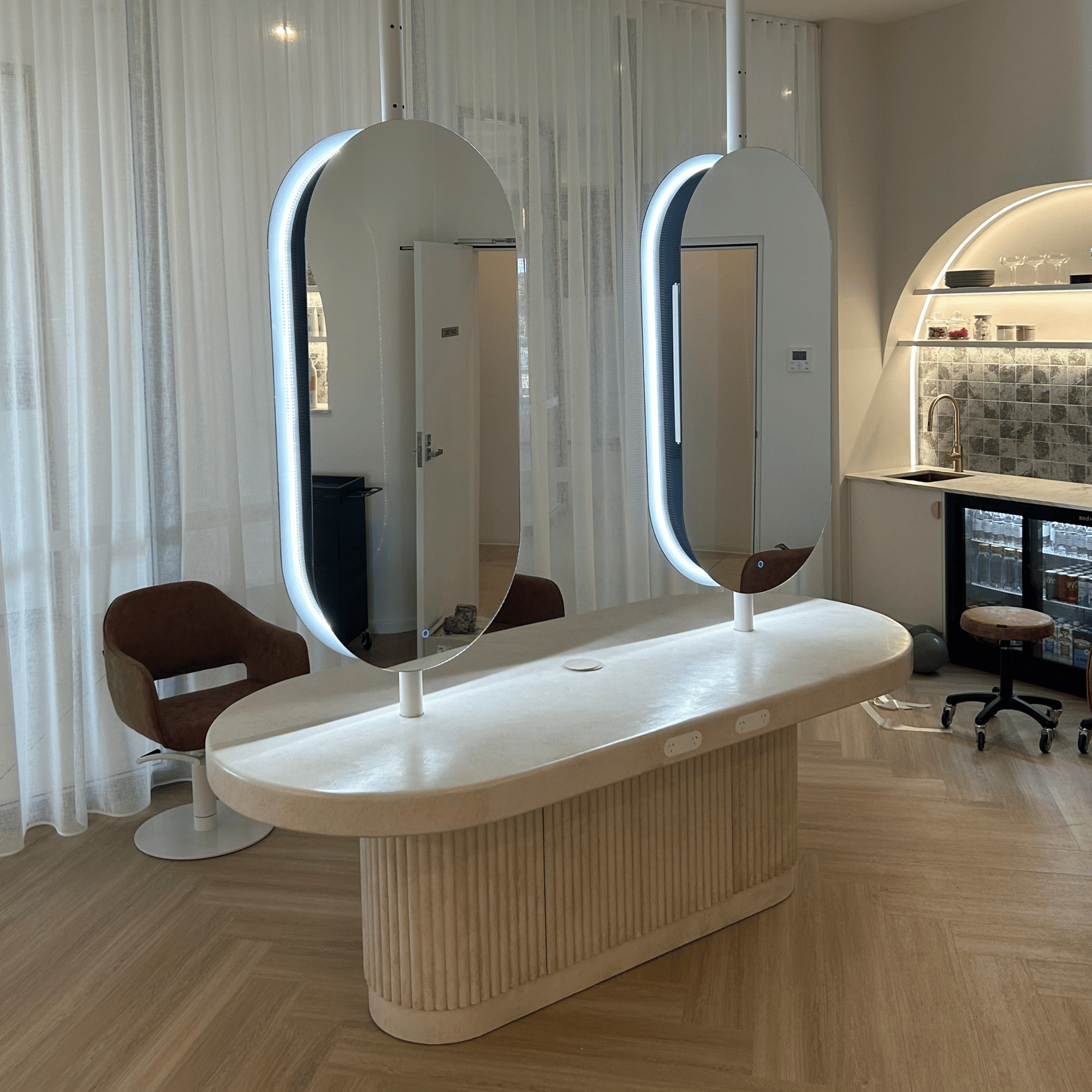 Modern vanity with two illuminated mirrors in a room with wooden flooring and a kitchen in the background.